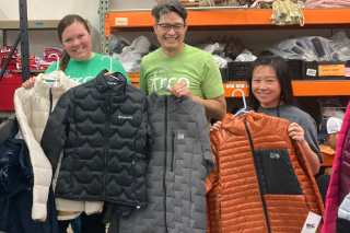 Three people holding up winter jackets with shelves of stocked good behind them